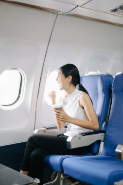 Çekici Asyalı kadın uçak yolcusu kahve içerken rahat koltukta oturuyor.