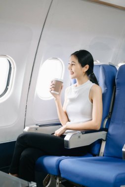 Çekici Asyalı kadın uçak yolcusu kahve içerken rahat koltukta oturuyor.