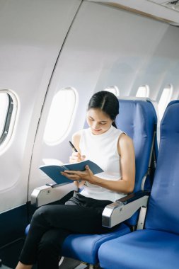 Çekici Asyalı kadın uçak yolcusu rahat koltukta oturuyor ve defterle çalışıyor. 