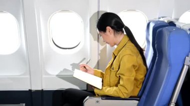 Resmi takım elbiseli Asyalı kadın yolcu rahat koltukta oturup not defterine notlar yazıyor. 