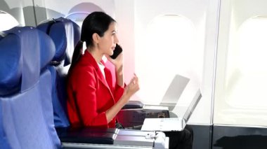 Asyalı kadın uçak yolcusu resmi bir takım elbise giyiyor rahat koltukta oturuyor dizüstü bilgisayar kullanıyor ve akıllı telefondan konuşuyor. 