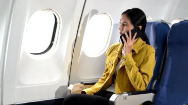 Resmi bir takım elbise giymiş, rahat koltukta oturan ve akıllı telefondan konuşan Asyalı kadın yolcu. 