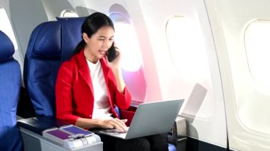 Asyalı kadın uçak yolcusu resmi bir takım elbise giyiyor rahat koltukta oturuyor dizüstü bilgisayar kullanıyor ve akıllı telefondan konuşuyor. 