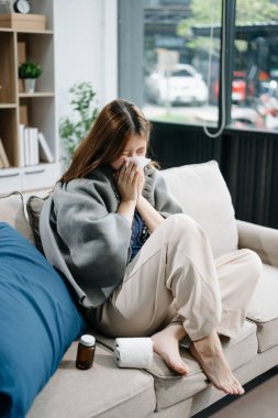 Kanepede battaniyeyle kaplanmış ve mendille hapşıran, grip belirtileri gösteren genç bir Asyalı kadın. Hastalık, sağlık ve iyileşme kavramı