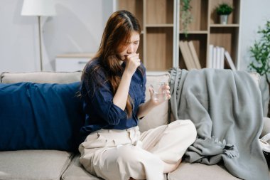 Genç Asyalı kadın grip belirtilerinden muzdarip ve kanepede bir bardak su tutuyor. Hastalık, sağlık ve iyileşme kavramı. Evde kal, sağlıklı kal.
