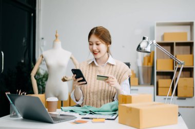 Genç iş kadını, akıllı telefon ve dizüstü bilgisayar kullanarak, müşterilere teslimat için paket kutuları hazırlıyor ve online siparişleri kontrol ediyor.