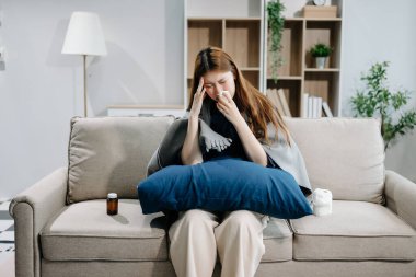 Kanepede battaniyeyle kaplanmış ve mendille hapşıran, grip belirtileri gösteren genç bir Asyalı kadın. Hastalık, sağlık ve iyileşme kavramı