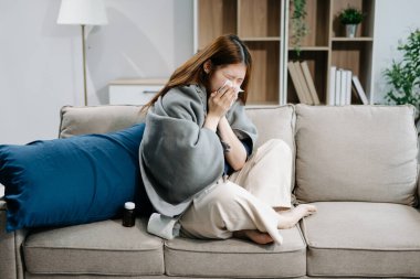Kanepede battaniyeyle kaplanmış ve mendille hapşıran, grip belirtileri gösteren genç bir Asyalı kadın. Hastalık, sağlık ve iyileşme kavramı
