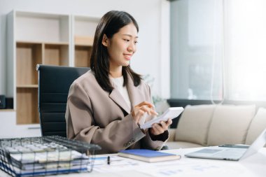 Genç ve güzel bir kadın çalışma yerinde otururken dizüstü bilgisayar ve hesap makinesi kullanıyor. İşine odaklan..