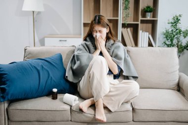 Kanepede battaniyeyle kaplanmış ve mendille hapşıran, grip belirtileri gösteren genç bir Asyalı kadın. Hastalık, sağlık ve iyileşme kavramı