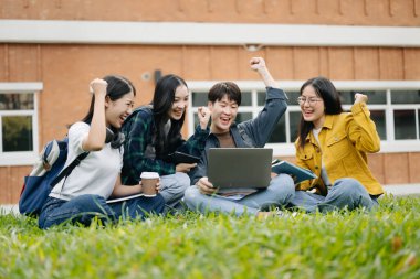 Genç Asyalı üniversite öğrencileri sabah kampüs parkında çimlerin üzerinde otururken ders çalışıyorlar. Birlikte vakit geçirmek, dizüstü bilgisayar ve dijital tablet kullanmak.