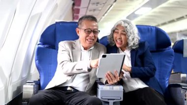 Tablet kullanarak uçakla seyahat eden ve pencereyi gösteren yaşlı Asyalı çift. 