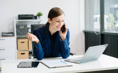 Dizüstü bilgisayarda çalışan ve yaratıcı ofiste akıllı telefonla konuşan kendine güvenen bir iş kadını.