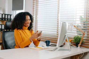 Ofiste çalışan Afro-Amerikan genç kadın akıllı telefon ve bilgisayar kullanıyor. 