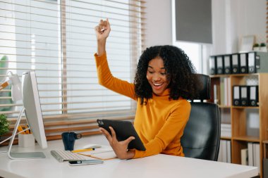 Mutlu Afro-Amerikan kadın ofiste tablet ve bilgisayar kullanarak çalışıyor.