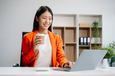 İş kadını laptop kullanıyor, modern ofiste ofis masasında kahve içiyor. 