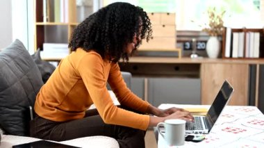 Genç bir kadın evdeki dizüstü bilgisayarını kullanarak rahat koltukta dinleniyor.