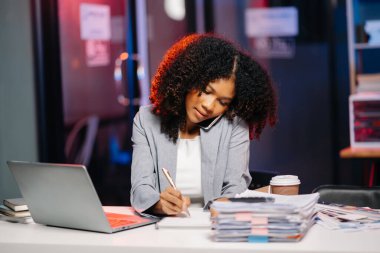 İş kadını kahve içiyor. Fazla mesai yapmak için enerji topluyor. Masa başında oturup, lamba ışığında fazla mesai yapıyor.