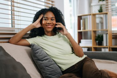 Mutlu kıvırcık Afrikalı kadın telefonda evdeki kanepede konuşuyor.