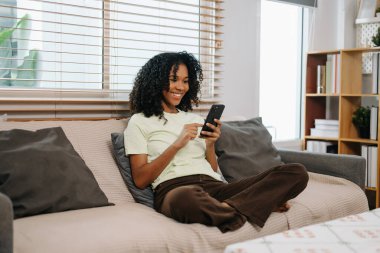 Genç kıvırcık Afrikalı kadın evdeki kanepede akıllı telefon kullanıyor.