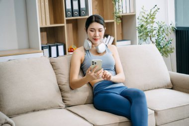 Genç Asyalı kadın spor kıyafetleriyle kanepede akıllı telefon kullanıyor. Evde otururken kulaklığı boynunda.