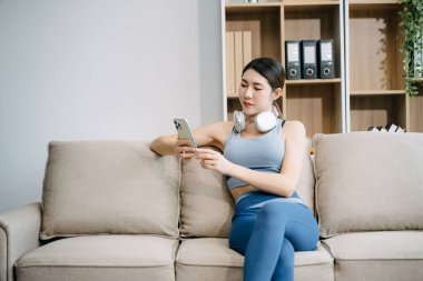 Genç Asyalı kadın spor kıyafetleriyle kanepede akıllı telefon kullanıyor. Evde otururken kulaklığı boynunda.