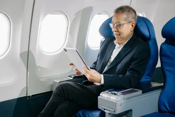 stock image Asian mature businessman wearing eyeglasses sitting in an airplane seat and using tablet 