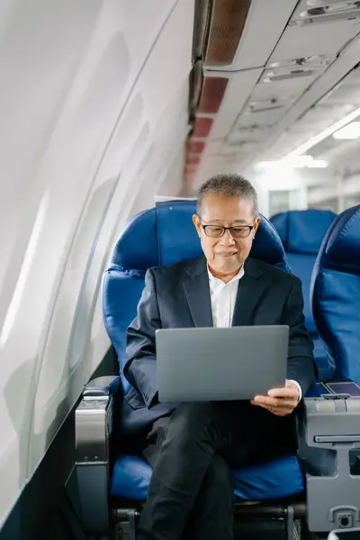 stock image Asian mature businessman wearing eyeglasses sitting in an airplane seat and using laptop 