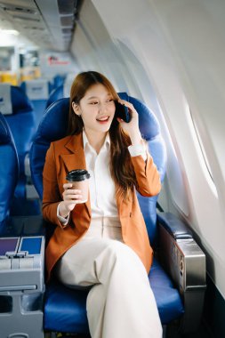 Başarılı bir Asyalı iş kadınının portresi resmi bir takım elbiseyle birinci sınıfta oturmuş akıllı telefon kullanıyor ve kahve içiyor. 
