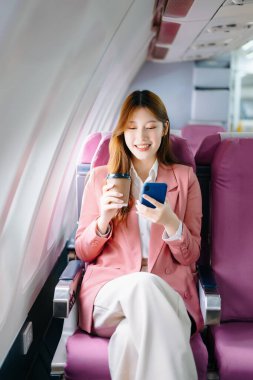Başarılı bir Asyalı iş kadınının portresi. Pembe ceketli, birinci sınıfta oturmuş akıllı telefon kullanıyor ve kahve içiyor. 