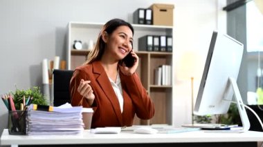İş kadını masada oturuyor, ofiste çalışıyor ve akıllı telefonla konuşurken bilgisayar kullanıyor. 