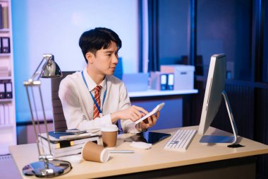 Asyalı işadamı fazla mesai yapmak için kahve içiyor. Masada oturup, lamba ışığında fazla mesai yapıyor.. 