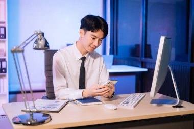 Fazla mesai konsepti, yakışıklı Asyalı iş adamı fazla mesai yapıyor bilgisayar, akıllı telefon kullanıyor ve lamba ışığında fazla mesai yapıyor.. 