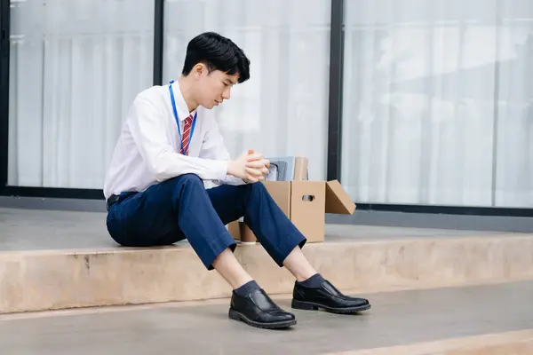 stock image Asian businessman sitting outside the office with a box of belongings, reflecting on job loss. 