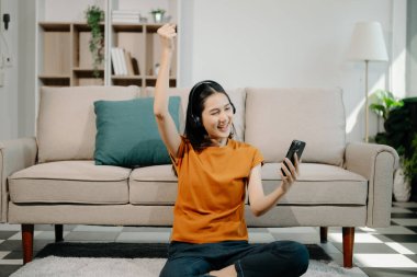 Genç Asyalı kadın evdeki kanepenin yanında otururken akıllı telefon ve kulaklık kullanıyor. 