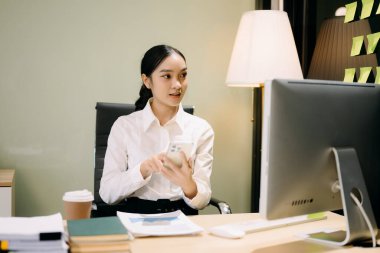 Asyalı iş kadını geceleri modern ofiste bilgisayar ve akıllı telefon kullanıyor.  