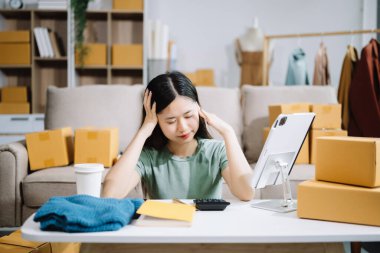 Young Asian female business owner prepare parcel boxes, woman is stressed, bored, and overthinking from working and standing check online orders for deliver to customer. clipart