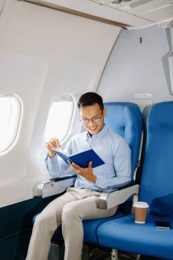 Sırıtkan Asyalı işadamı uçak koltuğunda kitap okuyor.  