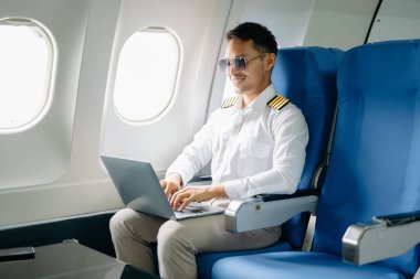 Gülümseyen Asyalı üniformalı erkek pilot yolcu koltuğuna yaslanıyor ve dizüstü bilgisayar kullanıyor. 