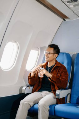 Asyalı bir adam uçak koltuğunda kahve içiyor ve pencereden dışarı bakıyor..
