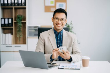 gülümseyen Asyalı işadamı ofiste çalışırken akıllı telefon ve laptop kullanıyor..
