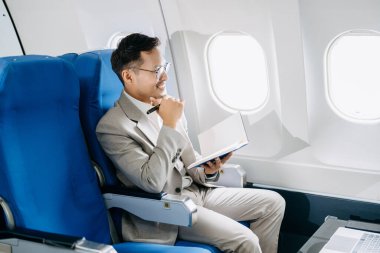 Gülümseyen Asyalı işadamının yan görünüşü, bir uçak koltuğunda çalışırken, defterine notlar yazarken.  