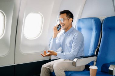 Asyalı erkek yolcu rahat koltukta oturup telefonla konuşuyor ve gülümsüyor. 