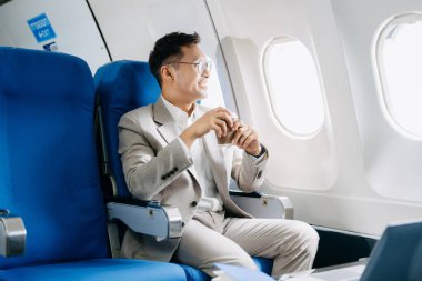 Asyalı işadamı uçak koltuğunda kahve içiyor ve pencereden dışarı bakıyor..
