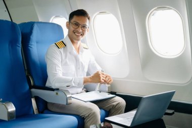 Gülümseyen Asyalı erkek pilot yolcu koltuğunda uçuş planlarını kontrol ediyor ve dizüstü bilgisayar kullanıyor. 