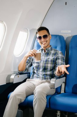 Güneş gözlüklü Asyalı bir adam uçak koltuğunda kahve içiyor, gülümsüyor ve kameraya bakıyor. 