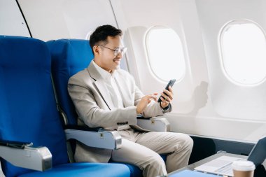 Sırıtkan Asyalı işadamı akıllı telefon ve dizüstü bilgisayar kullanarak uçak koltuğunda çalışıyor. 
