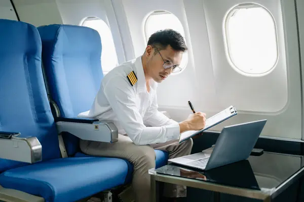 stock image Asian Confident male pilot in uniform at passenger seat of airplane checking flight plan and using laptop 