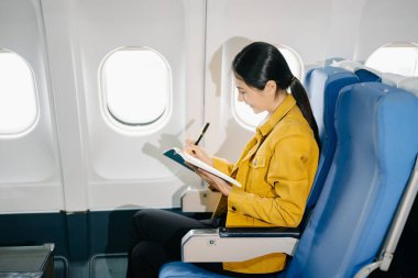 Çekici Asyalı kadın uçak yolcusu rahat koltukta oturuyor ve deftere yazıyor. 