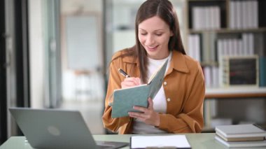  Güzel bir iş kadını dizüstü bilgisayarda çalışıyor ve ofisteki masada notlar alıyor.